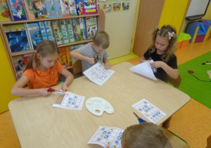 Czworo dzieci siedzi przy stoliku, w ręce trzymają nożyczki i wycinają z kartki kolorowe znaki drogowe.